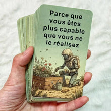 Une main tenant un jeu de 56 cartes oracle en français illustré avec un astronaute et des plantes vertes