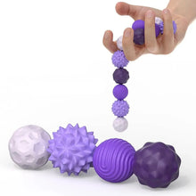 Une main tient une chaîne de balles sensorielles magnétiques en silicone violet et blanc avec textures variées
