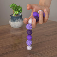 Une main tient un lot de 8 balles sensorielles magnétiques en silicone colorées et texturées violettes et blanches