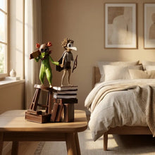 Deux figurines fantaisistes little nightmares iii, une verte avec bois orange et lunettes tenant un livre, l’autre avec cape et arc, sur des livres empilés