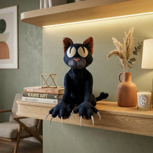 Peluche chat monstre noir avec yeux blancs et nez rose assis sur une étagère en bois
