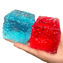 Une main tient deux cubes squishy sensoriel translucides en résine brillante bleue et rouge texturée et cristalline